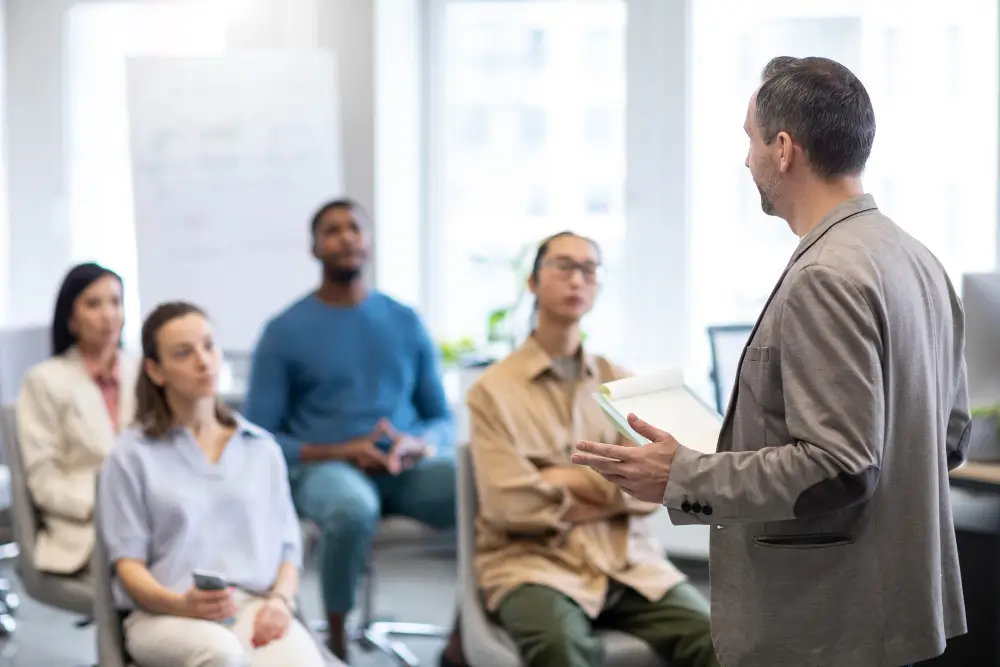 Speaker presenting car buyer consulting guidance to a group, explaining how to avoid dealership scams, hidden fees, and save money.
